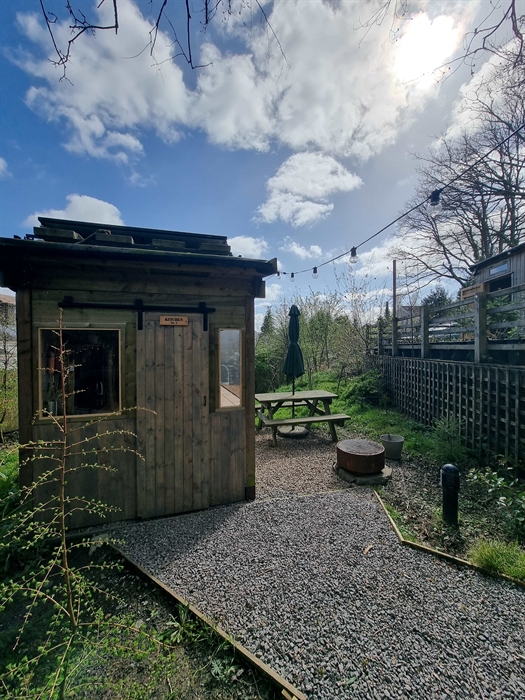 A cabin with a picnic table and firepit surrounded by greenery and a blue cloudy sky