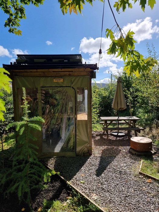 an outdoor kitchen next to a picnic table and a firepit surrounded by green trees and plants and a sunny blue sky