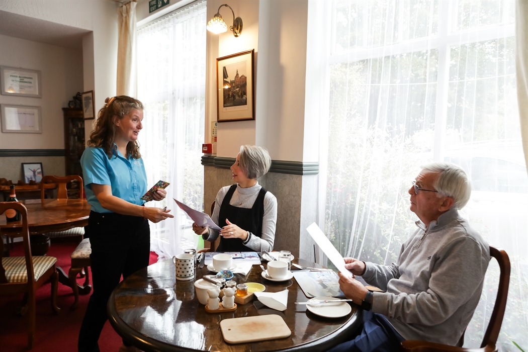 In this image Tracey is wearing a blue shirt with the restaurant windows letting all the light in onto 2 guests sitting at the table ordering their br