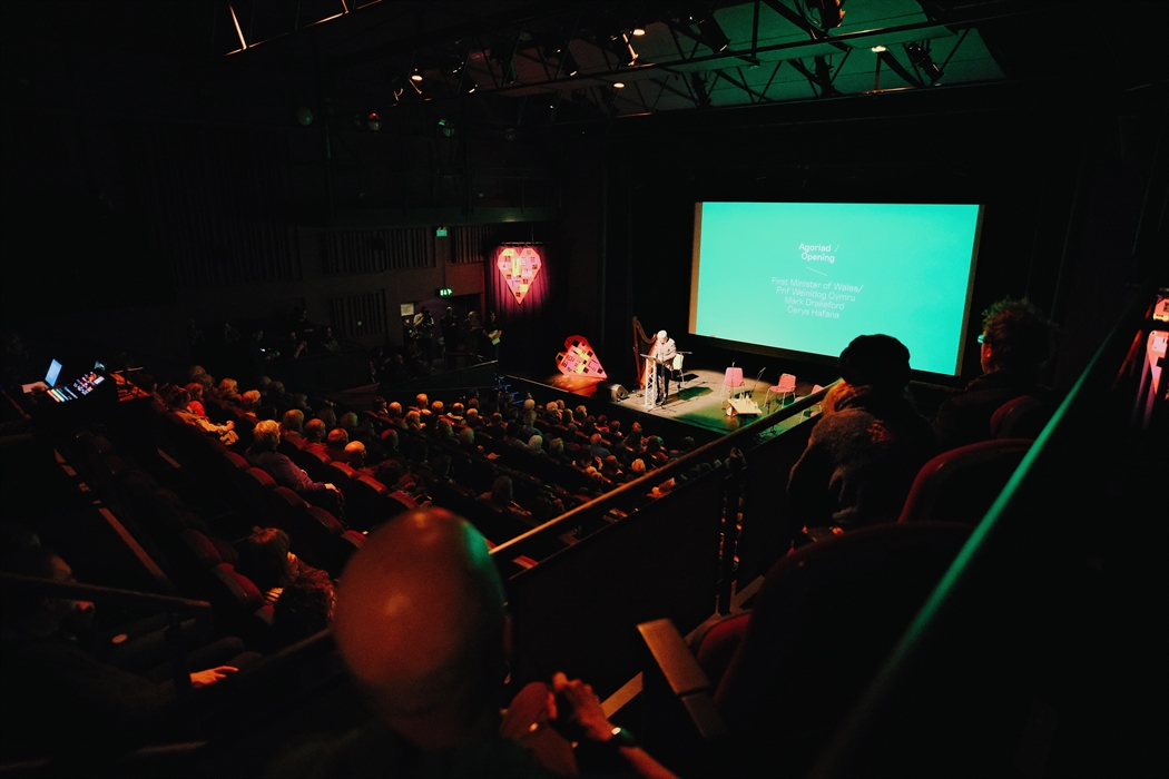 A view of an auditorium full with dimly audience members and a man standing on the stage, lit up and with a turquoise screen behind him. in the foregr
