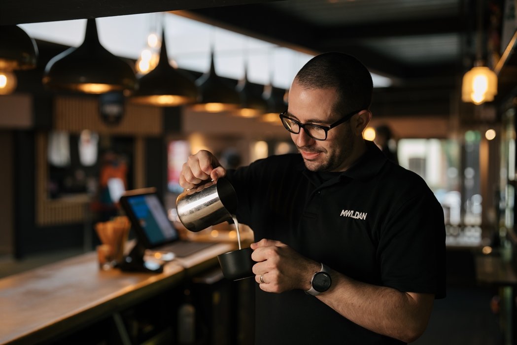 A man stands behind a bar as he pours milk out of a jug into a coffee cup. He wears a watch, glasses and a dark t-shirt with a logo that reads 'MWLDAN
