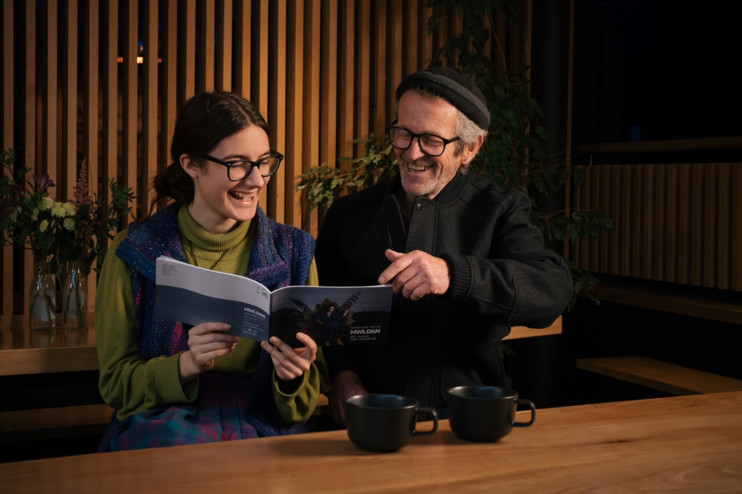 A young woman with dark hair, glasses, a green polo neck and a blue waistcoat sits next to a man with a dark hat, glasses and dark jacket as they look