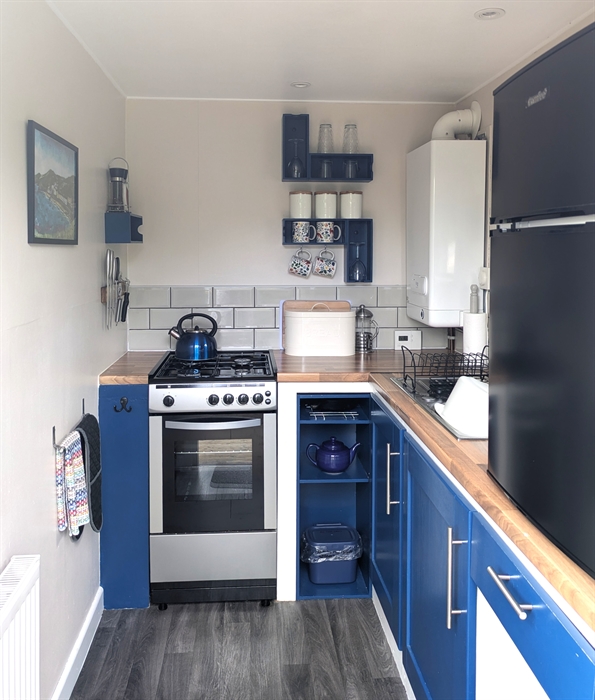 Blue cupboards and shelves. Gas cooker with hob top kettle, boiler, sink, and counter top fridge.