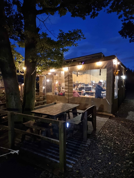its night time and in a wooden building, we can see people sat down. The building is under trees and lit up with string lights