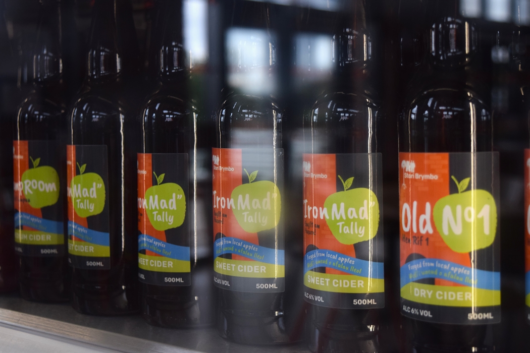 Rows of dark bottles in a fridge with labels on. The labels are for different types of cider. They have a picture of an old blast furnace in orange an