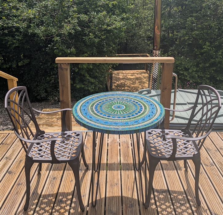 Handmade mosaic table in blue and green with metal dining armchairs on a wood deck with the hot tub, trees and lake in the background.