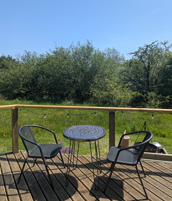 A handmade mosaic table in iridescent glass with 2 black woven chairs on the wood deck looking out to the meadow and the woodland behind.