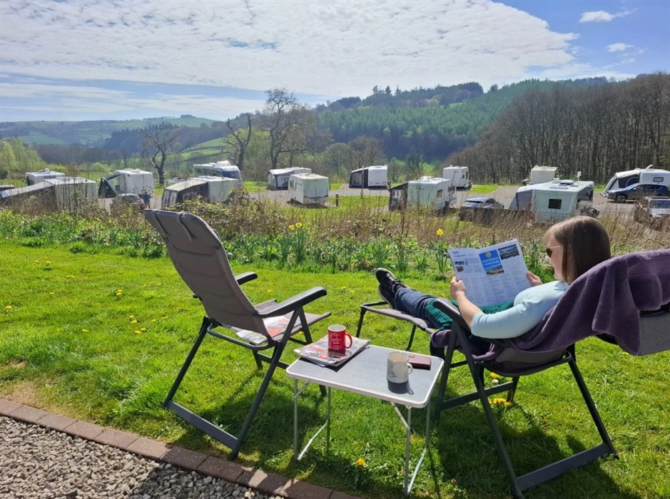 A photo of a woman relaxing on holiday, sat out on a sun lounger reading a magazine overlooking the Clywedog Valley.