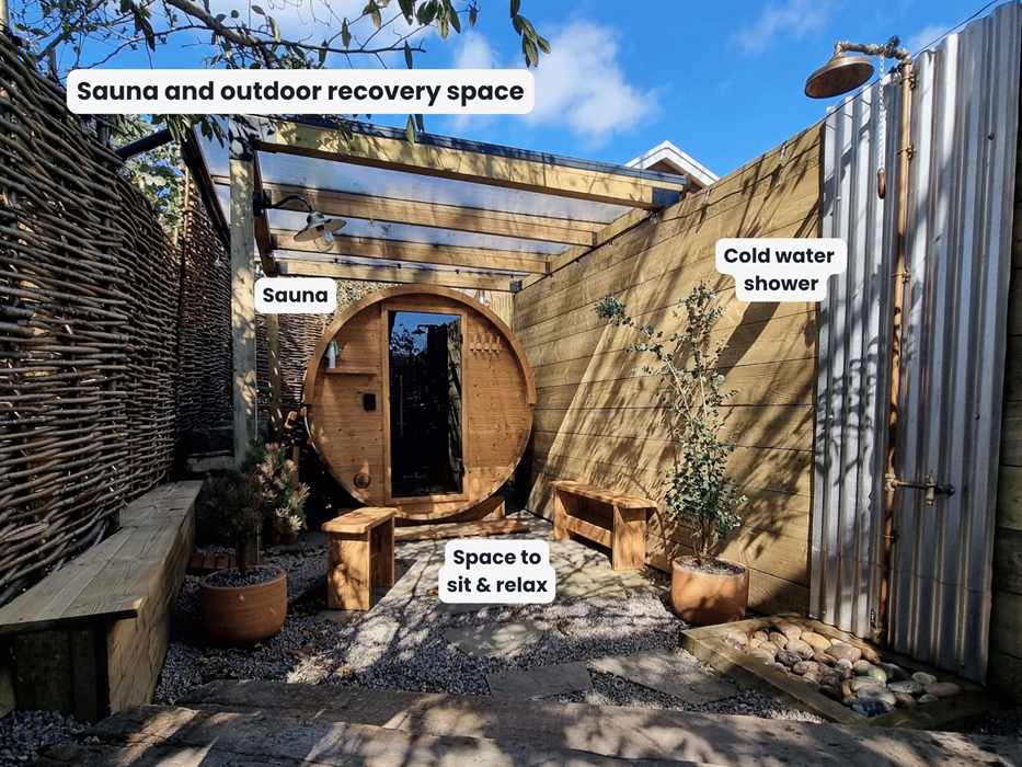 An outdoor, round wooden barrel sauna tucked away under the trees with an outdoor shower and benches for sitting on