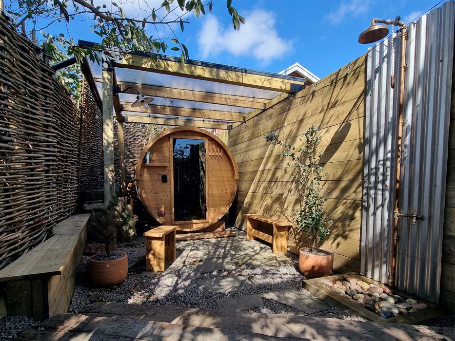 An outdoor, round wooden barrel sauna tucked away under the trees with an outdoor shower and benches for sitting on