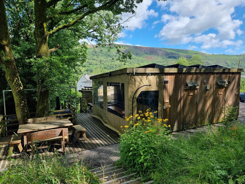 A wooden cabin with a picnic table surrounded by trees and a backdrop of a mountain and blue sky