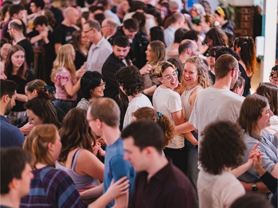 A large, joyful crowd of people dance closely together in a warmly lit room. Smiling faces and casual attire create a lively, festive atmosphere.