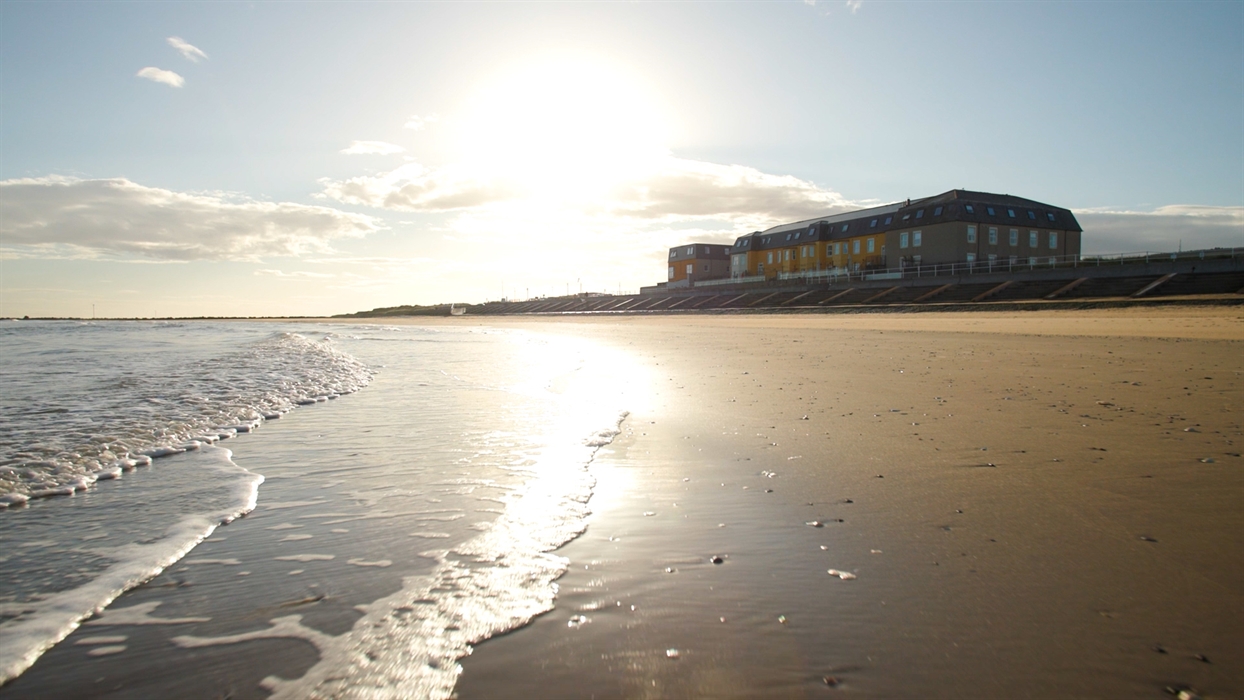 Looking up at The Beaches Hotel from Barkby Beach. Image shows the sand and the waves on the shore, and the hotel in the background.