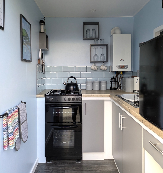 A gas stove with hob top kettle, boiler, sink and black counter top fridge. Tea,coffee,sugar canisters, coffee pot are on the work top.