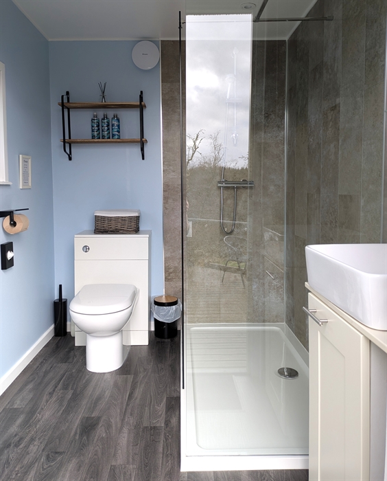 A glass shower enclosure, with brown marble Aquaboard, white toilet and handbasin. The shelf above the toilet holds Faith in Nature toiletries.