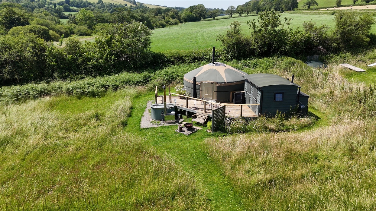 Green Yurt on a raised deck with dark green wood hut on right of photo, wood fired hot tub to left. Bench and fire pit to front surrounded by wild flo
