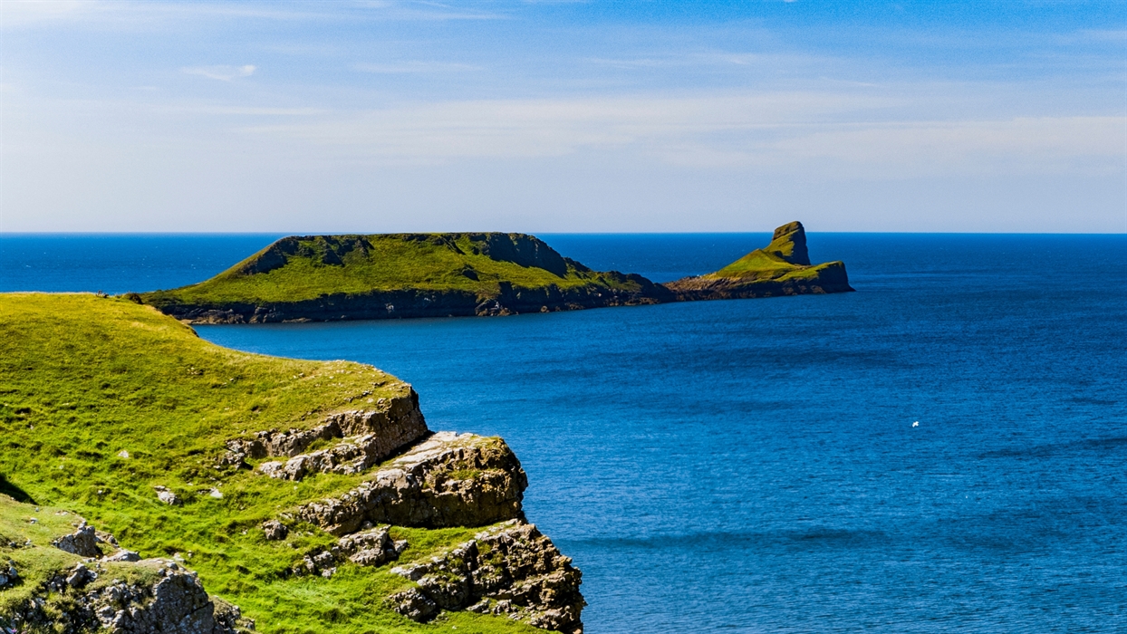 A bright, sunny day at the coast with green, grassy cliffs overlooking a vast, deep blue ocean. In the distance, two rocky islands rise from the sea,