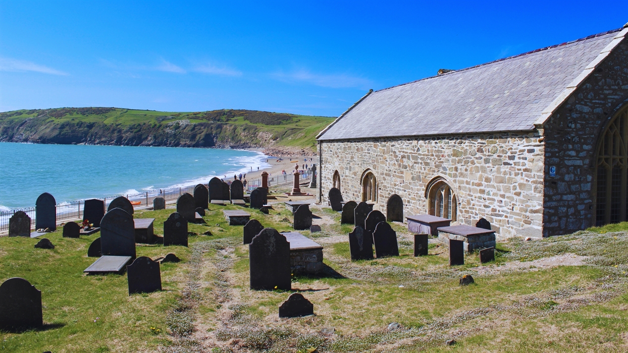 A stone church sits on a grassy hill overlooking a beach and the ocean, with a graveyard in the foreground.