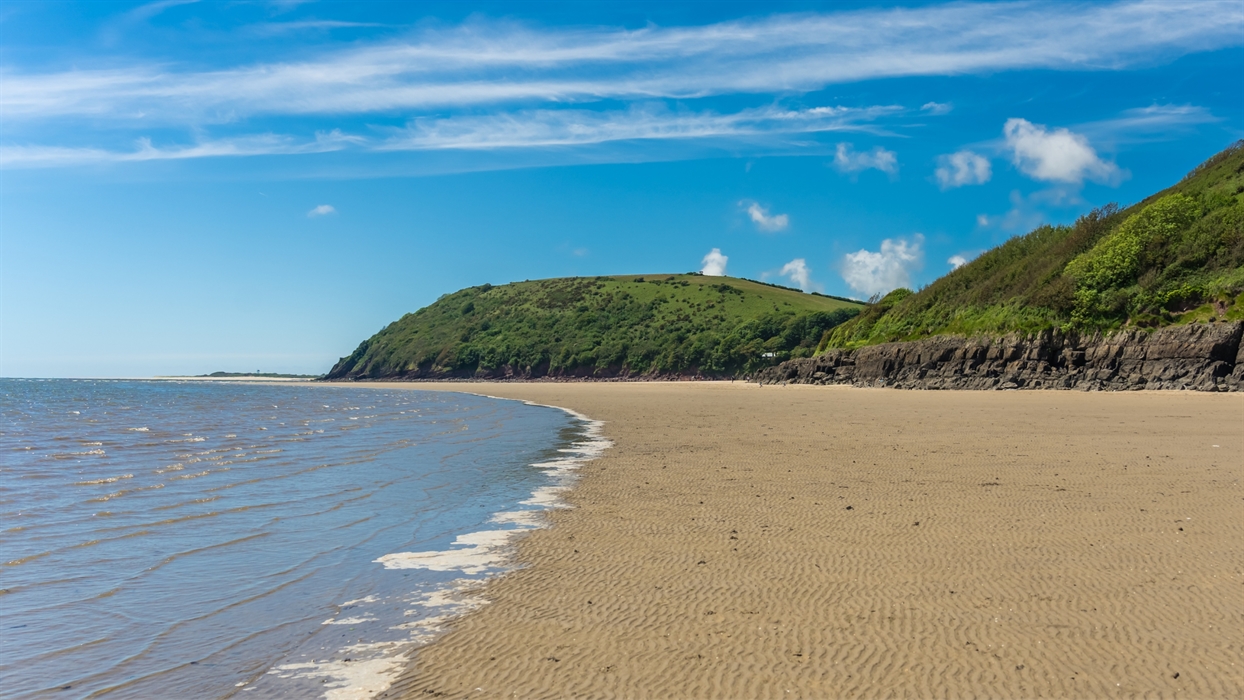 A sandy beach with gentle waves lapping the shore under a bright blue sky with wispy white clouds. Green, tree-covered hills rise from the background
