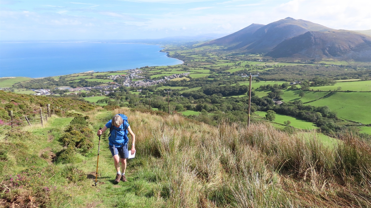 A hiker with a blue backpack and trekking poles walks down a grassy hill towards a coastal town with mountains in the background.