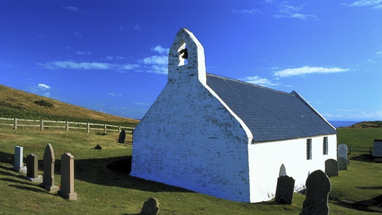 A small white church with a bell tower stands in a grassy graveyard under a bright blue sky with scattered clouds.