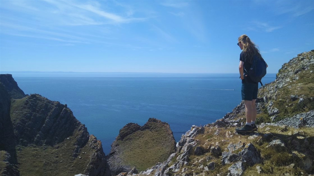 A person wearing a backpack stands on a rocky cliff overlooking a vast blue ocean under a clear sky.