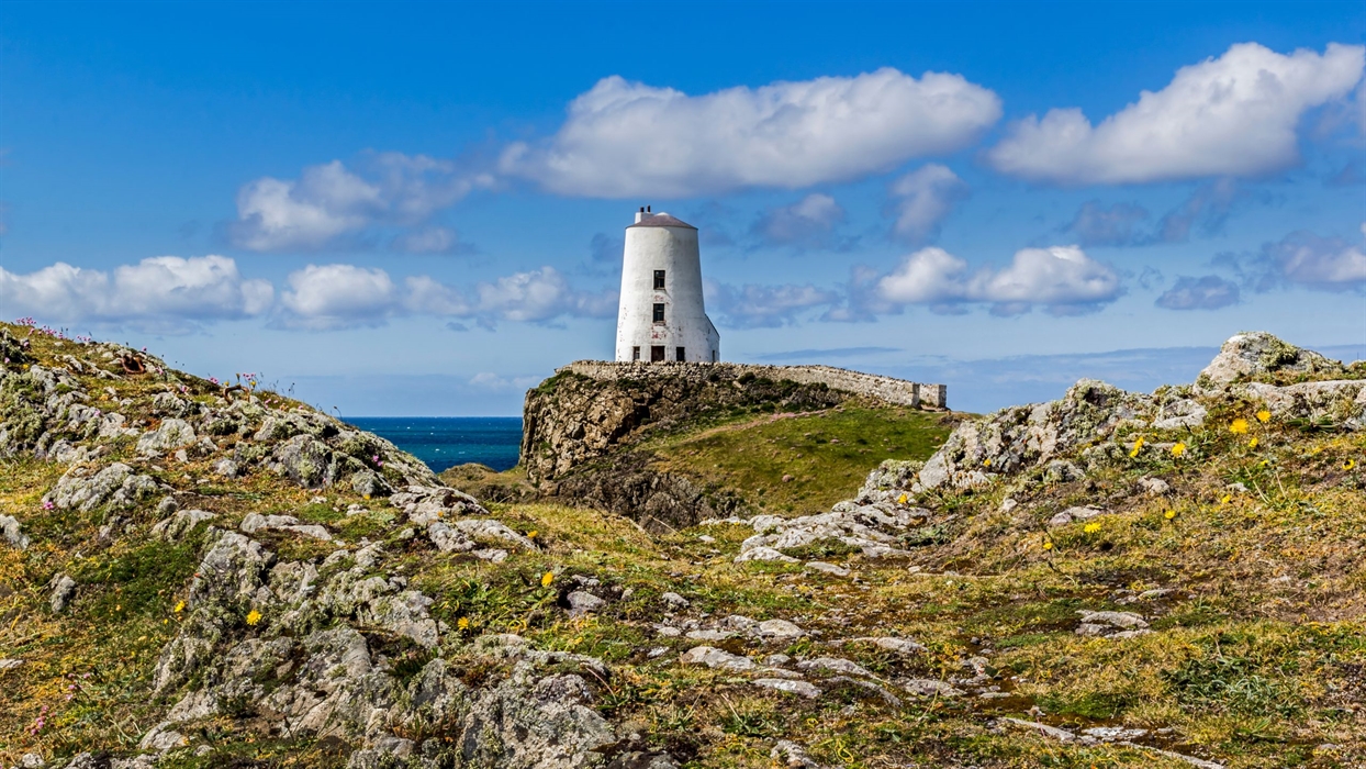 A white lighthouse stands on a rocky outcrop overlooking the sea under a blue sky with white clouds.