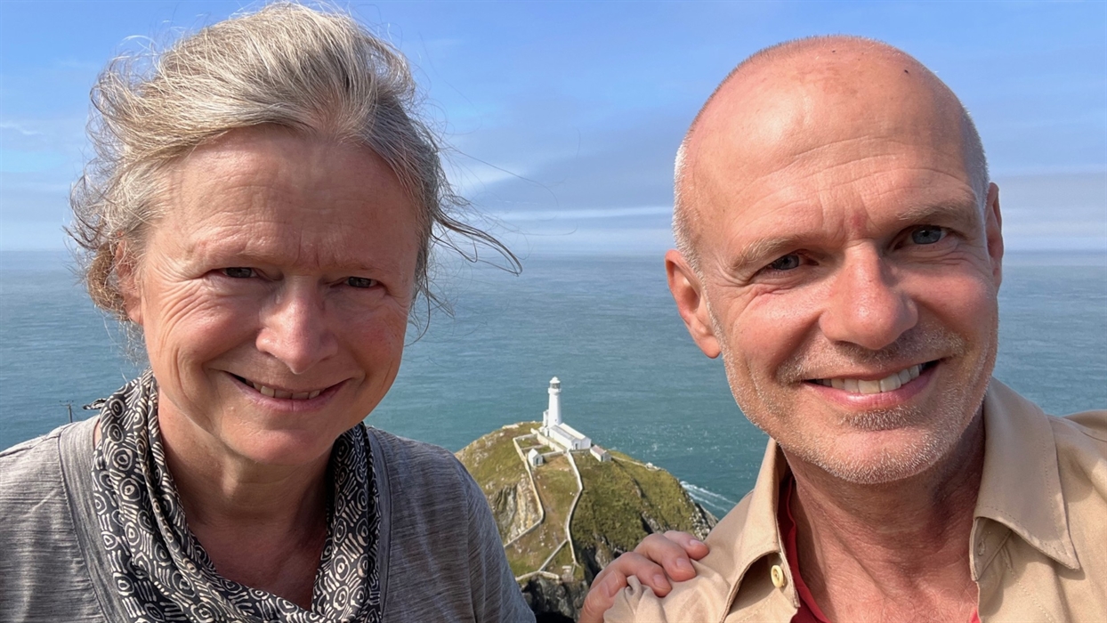 A close-up selfie of two people smiling with a lighthouse on a rocky outcrop in the background under a bright blue sky.