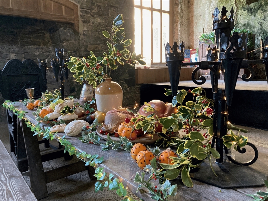A long wooden table laden with a festive spread of bread, oranges and a roasted fowl, decorated with holly branches and fairy lights.