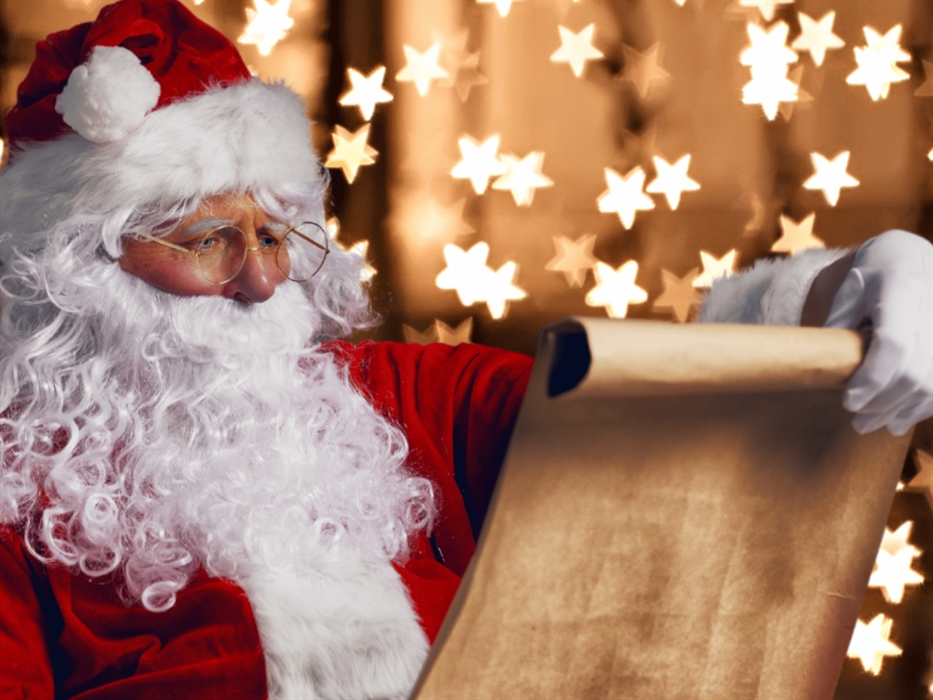 Santa Claus with a white beard and red suit is reading from a rolled-up parchment scroll, with blurred star-shaped Christmas lights in the background.