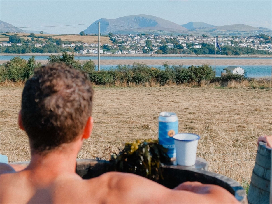 A summery image of a man bathing at Halen Môn's Wild Môn Seaweed Baths, in a barrel of seaweed with a Halen Môn Tide lager and a view of the mountains