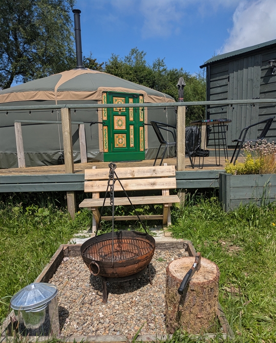 a steel fire pit with tripod in a pebbled enclosure in front of a wood bench, which is in front of a raised deck with a yurt and hut off.