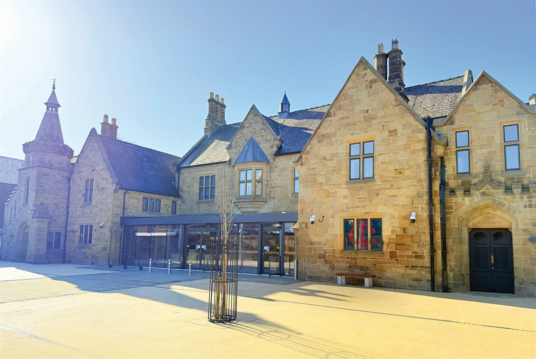 Photo of a historic stone building with chimneys, a modern glass front and a sand coloured forecourt, a small tree and bench, against a blue sky.