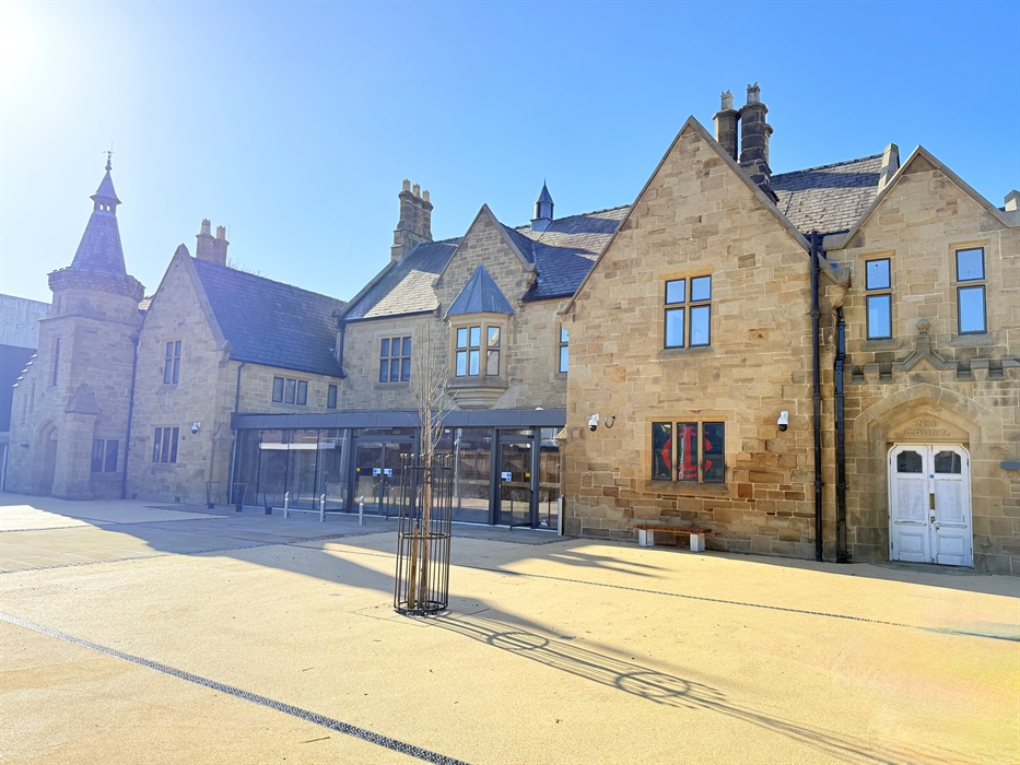 Photo of a stone building with chimneys, a glass front and a sand coloured forecourt, a small tree and bench, against a blue sky.