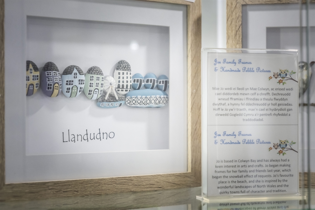 Handmade pebble art frame showing a Llandudno seaside scene alongside information about a local artist from North Wales.
