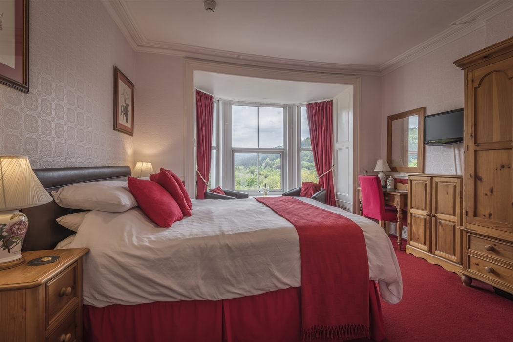 A cosy guest room with a double bed dressed in white linens and red accents. The room features wooden furniture, including bedside tables, a wardrobe,
