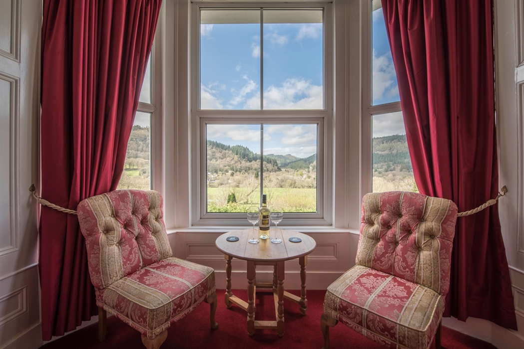 A pair of ornate red‑and‑gold upholstered chairs sit in front of a large window framed by deep red curtains. A small round table between them holds a