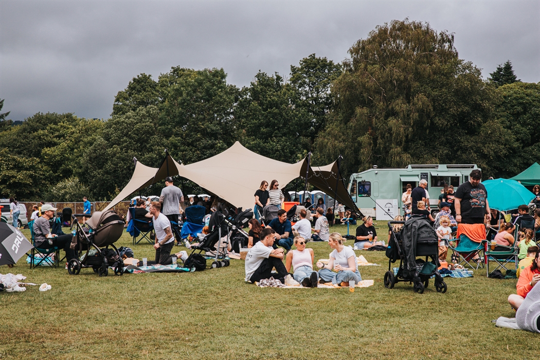 Outdoor food festival scene with families and groups of friends relaxing on grass in front of stretch tents and food trucks. People sit on picnic blan