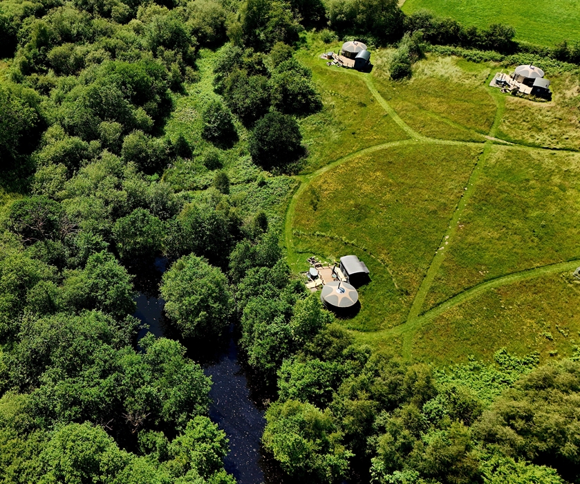 Bird's eye view of 3 yurts in a meadow, surrounded by woodland and with a small lake in top left.