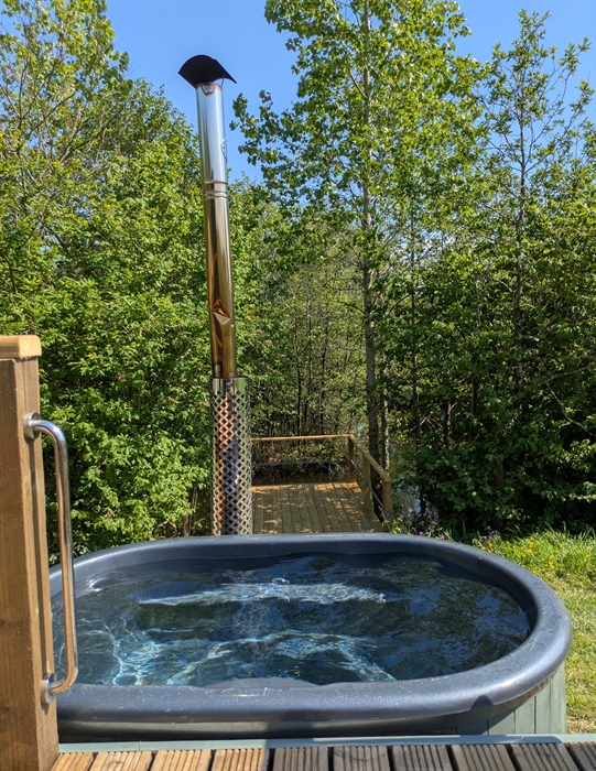 Grey fibreglass hot tub in wood casing next to the wood deck, with a metal chimney behind, overlooking the lake and trees in the background. There are