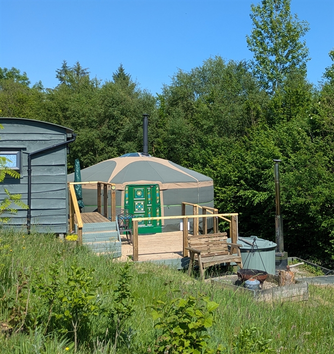 A green yurt with a bright green decorated door, on a wood deck with a dark green painted wood hut up some steps on the left of the image, a dark gree