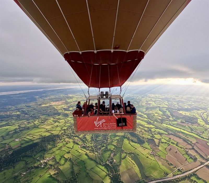 Passengers enjoying a Virgin Balloon Flights hot air balloon ride over the Welsh countryside, with patchwork fields and sunlit views below.