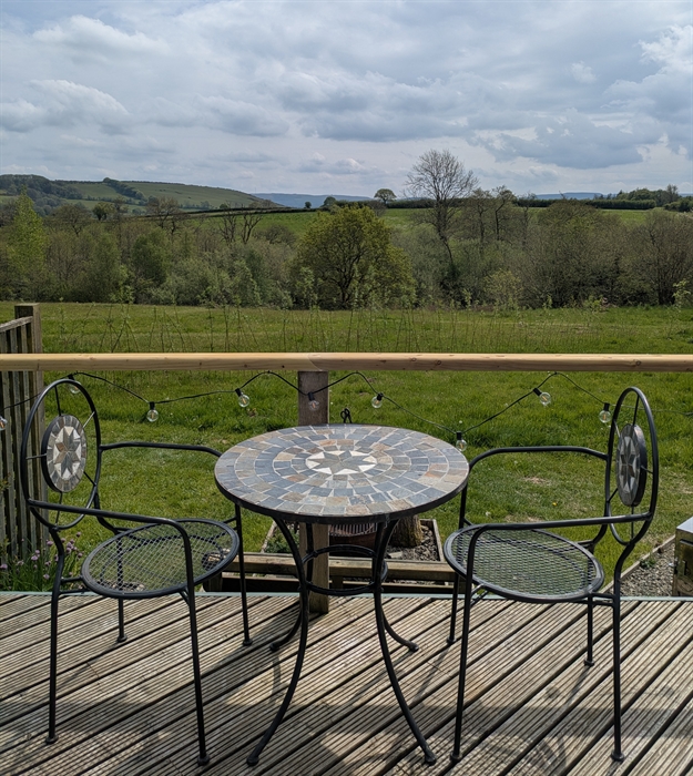 Metal and stone mosaic outdoor table and chairs on the wood deck with a view over the wildflower meadow to the trees round the lake and the hills beyo